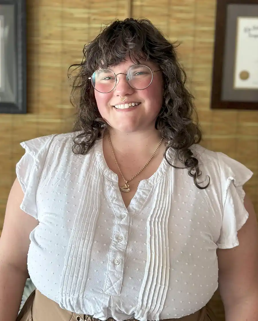 woman massage therapist in white shirt curly brown hair wearing glasses and smiling against a bamboo wall acupuncture office in key west