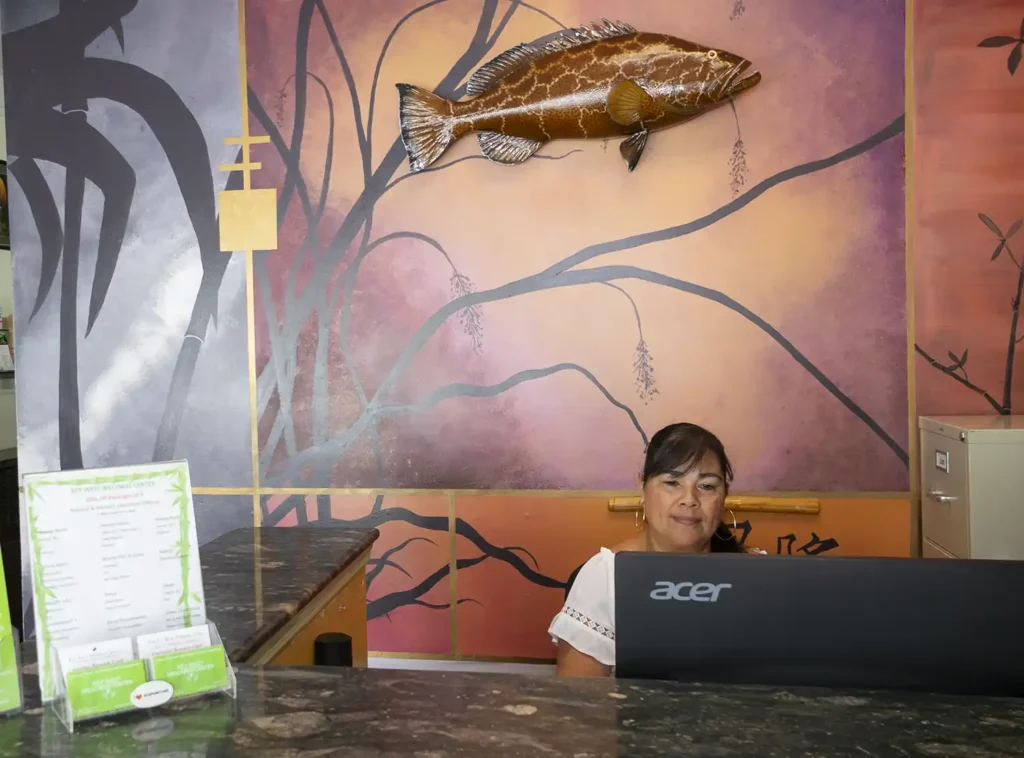 dark haired female office manager in front of a computer behind a desk and next to a painted wall of bamboo and a fish hanging in an acupuncturist office