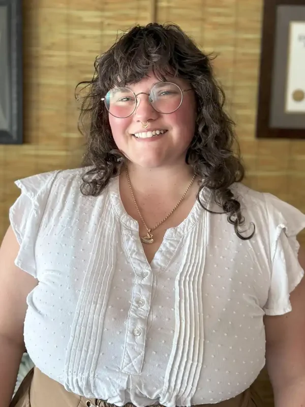 woman massage therapist in white shirt curly brown hair wearing glasses and smiling against a bamboo wall acupuncture office in key west